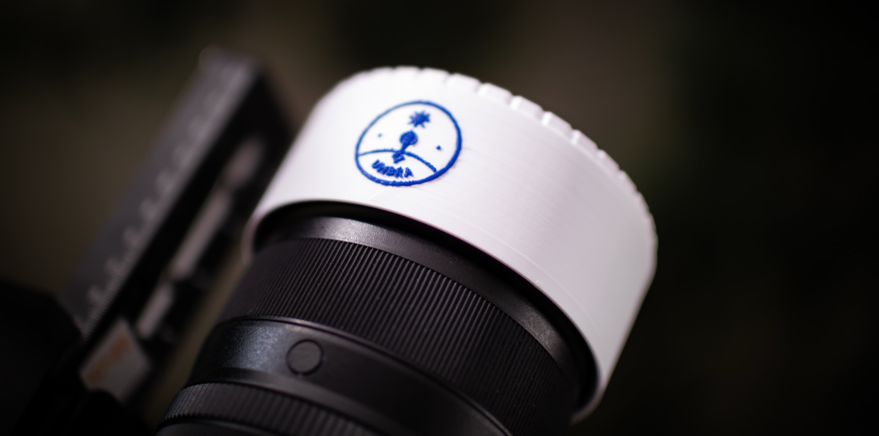 A white 3D-printed Umbra solar filter pushed onto the front of a camera lens, with the Umbra badge on the skirt.