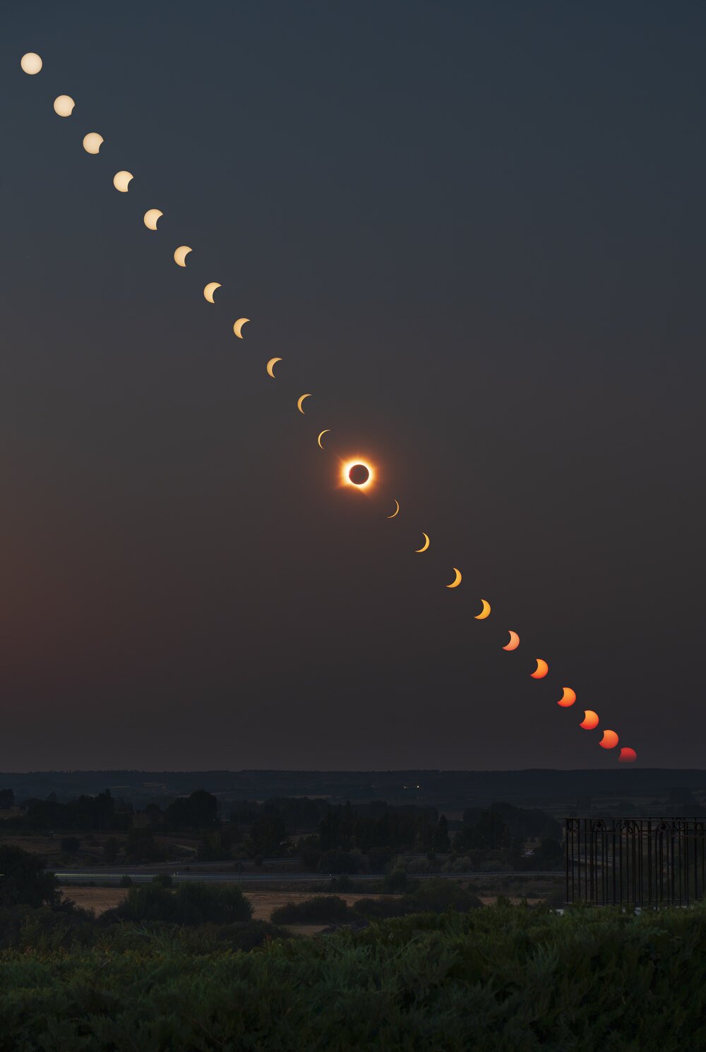 A composite of the whole eclipse: a diagonal arc of partial phases running down the sky to totality and out again, over a Spanish landscape at dusk.